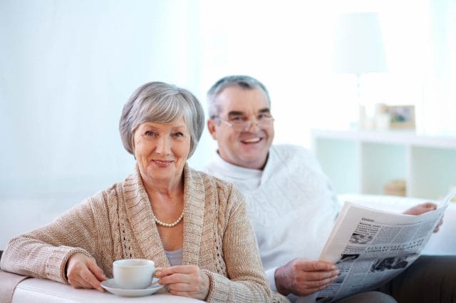 A mature woman with coffee cup and older man with newspaper sit on couch.