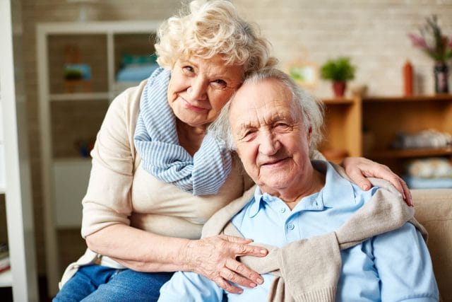 An older couple smiles and hug and sit on a couch in their home.