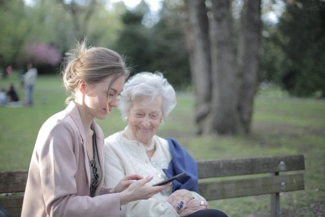 A young woman sits on a park bench with her grandmother and shows her a picture.