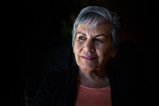 A portrait of an older woman in front of a dark background, looking off into the distance.