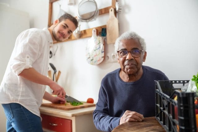 An older adult man sits at a kitchen table. A younger man stands behind him and prepares a meal on the kitchen counter.