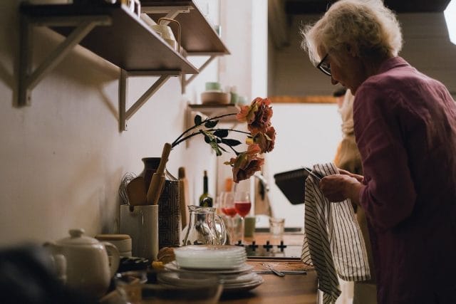 A picture of an older woman standing in a dim kitchen drying dishes.