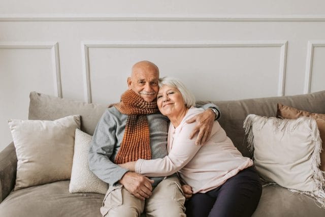 An older couple sit on a couch at home and smile to the camera while hugging.