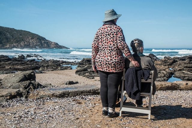Two woman look at the coastline on a rocky beach. One woman sits in a chair and the other stands next to her.