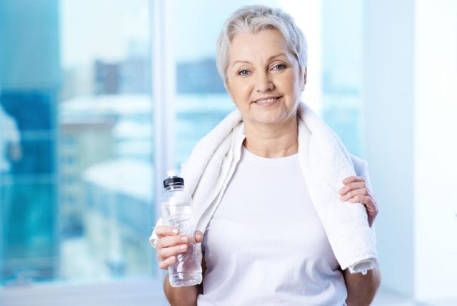A senior woman smiles at a camera, wearing a tshirt and towel around her neck. She's holding a water bottle.