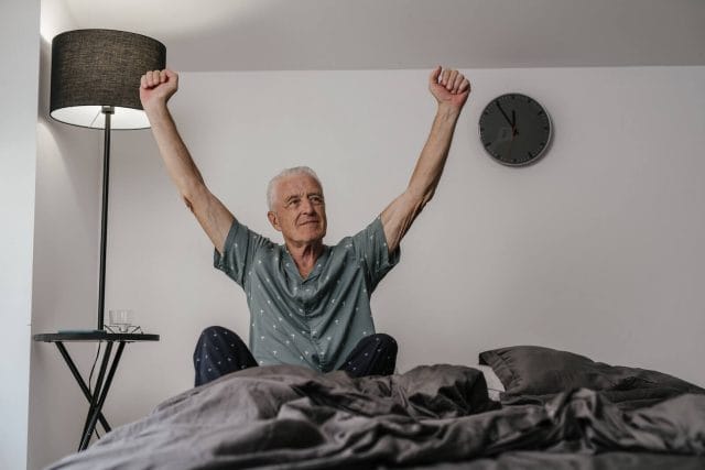 an older adult man sits up in bed and reaches his arms over his head.
