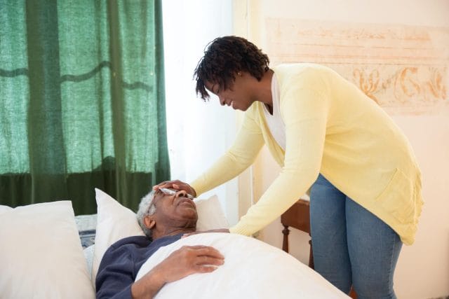 A young woman feels the forehead of her older loved one as he lays in bed.