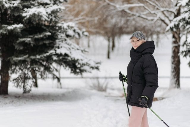 A senior woman wears a heavy winter jacket and uses walking poles to walk outside on a snowy day.