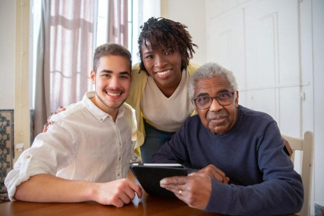 Two young adults and a senior man smile at the camera. The man holds a tablet.