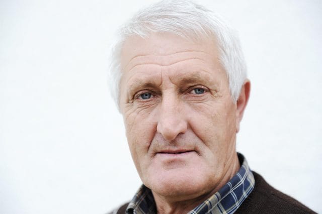 A close-up of an older adult man with white hair and a collared shirt and sweater.