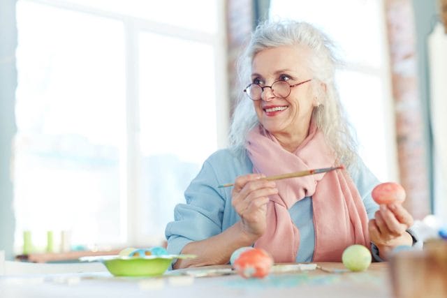 A senior woman holds a paint brush and is painting an art project. She wears glasses and a scarf while smiling.