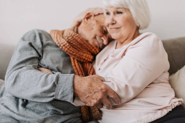 A senior woman embraces a senior man as he rests his head on her shoulder.
