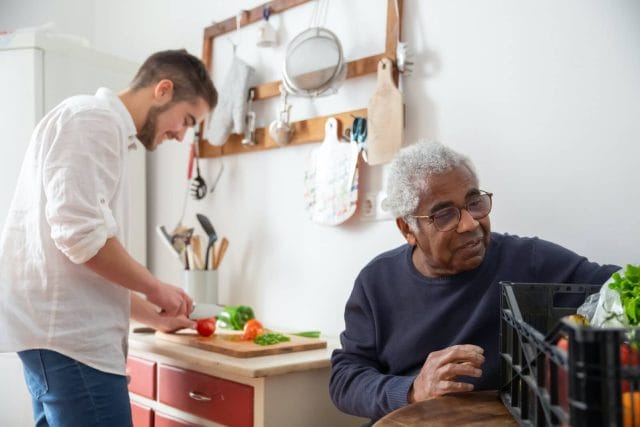 A young man prepares a meal for a senior man who sits at a table next to the counter.