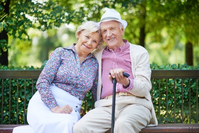 A happy senior couple sit on a park bench and smile.