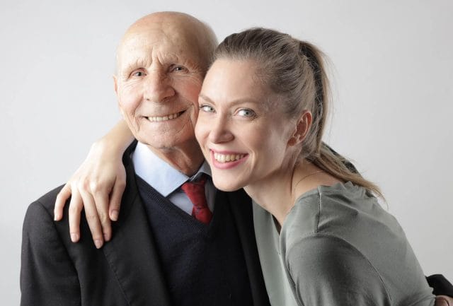 A young woman smiles and wraps her arm around a senior man wearing a suit. He smiles at the camera.
