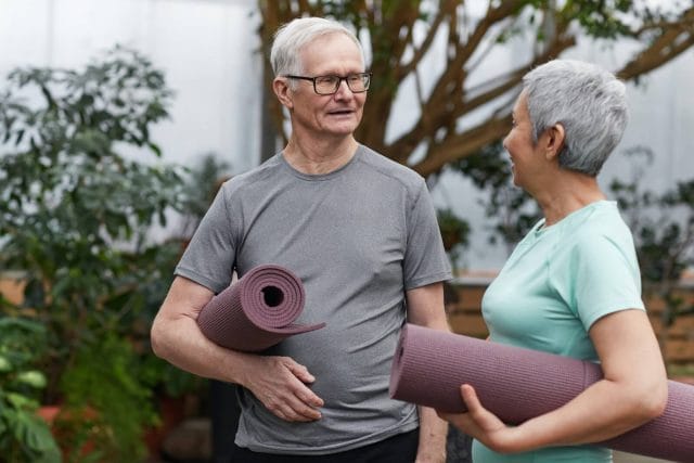 An older adult man and woman stand in conversation with each other, each holding a rolled up yoga mat.