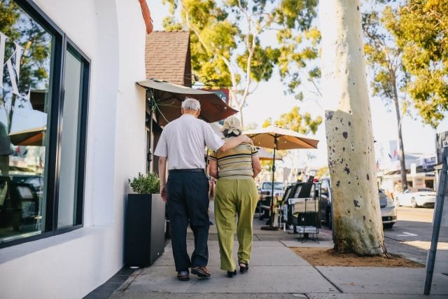 Two older adults walk on a sidewalk on a sunny day. One has his arm around the other's shoulder.