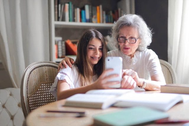 An older adult woman sits at a table with her granddaughter. They smile and look at a smartphone.