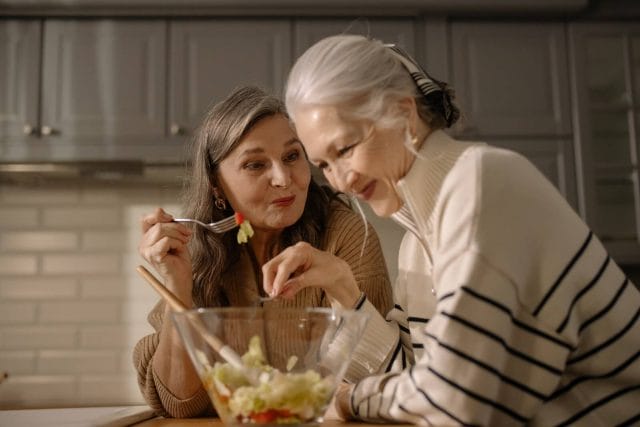 Two older adult women laugh and eat healthy snacks.