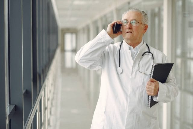 an older adult doctor holds a notebook and takes a phone call on his smartphone