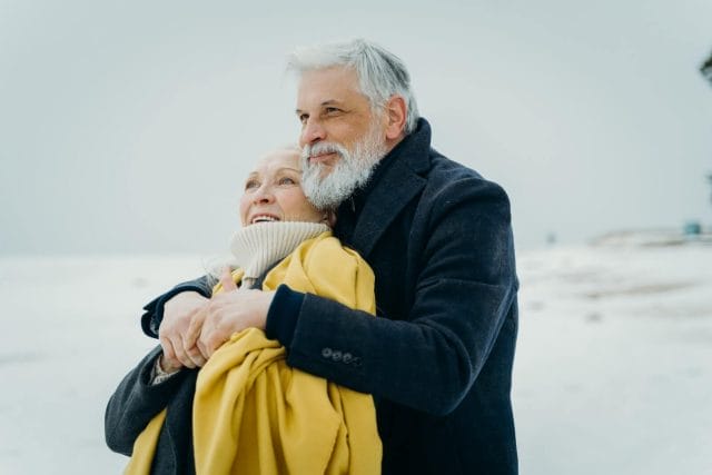 An older adult couple hugs each other and looks off into the distance.