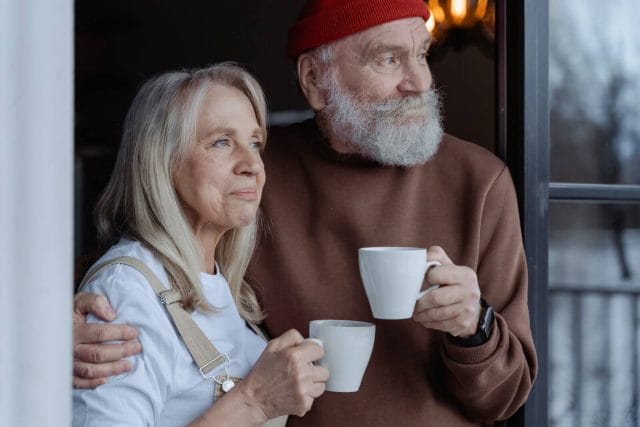 A man and woman stand near a window and look out of it, holding coffee mugs in their hands.