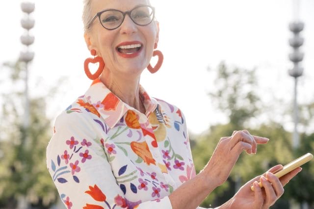 A smiling older adult woman wears a vibrant shirt, heart-shaped earrings, and glasses. She looks at the camera and holds a smartphone in her hand.