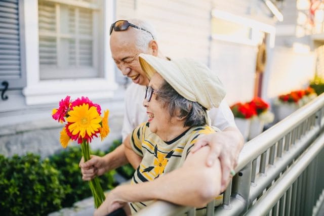 An older adult couple smiles and laughs as they lean against a fence. The woman holds a bouquet of flowers.