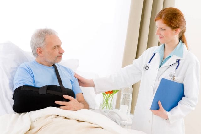 A patient with a sling on his arm lies in a hospital bed. His doctor holds a file and comforts him.