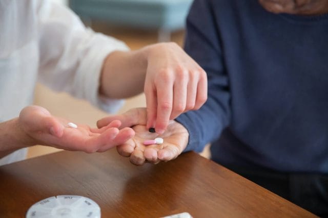A close-up image of a nurse's hands placing medication into a patient's hands. A medication organizer is on the table.