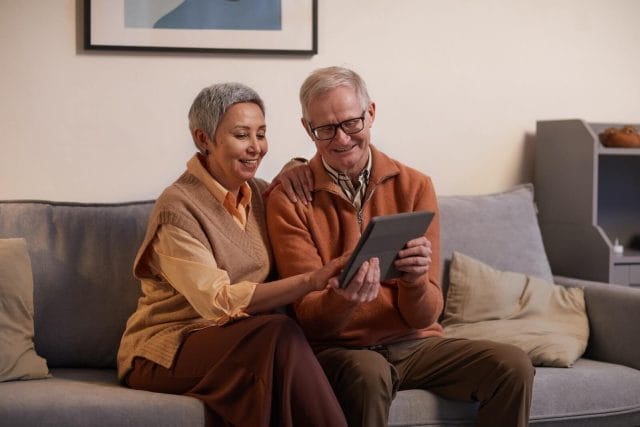 An older adult man and woman sit on a couch and look at a tablet together. They are reviewing information.