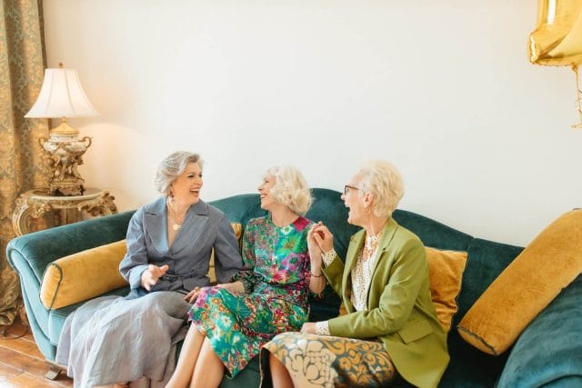Three older adult women sit on a green velvet couch and laugh together.