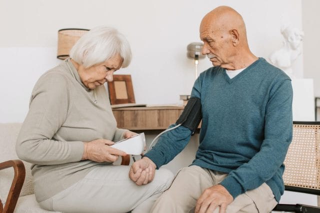 A senior woman checks an older adult mean's blood pressure using a blood pressure monitor in their home. They appear concerned.