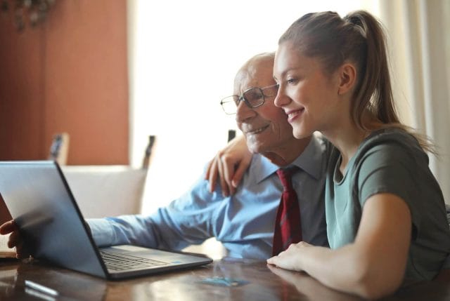 A young woman and a senior man sit at a desk and look at information on a laptop computer.
