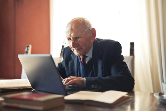 Older adult man wears professional clothing and glasses. He sits at a desk and works on a laptop computer.