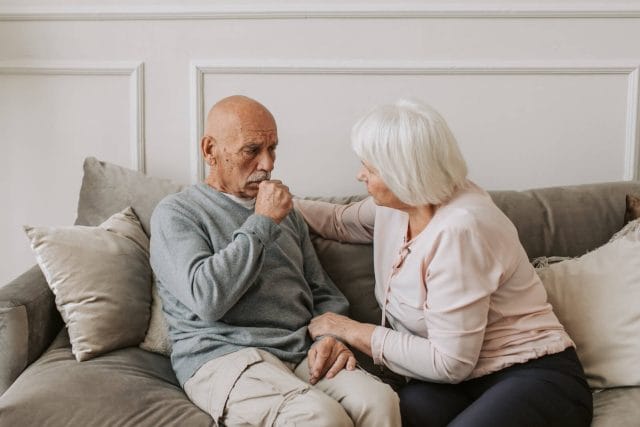 An older adult woman and man sit on a couch. The man appears to cough or is in distress. The woman looks at him, concerned.