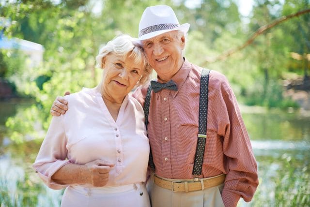 A smiling older adult couple stands outside and smile at the camera, their arms around each other.