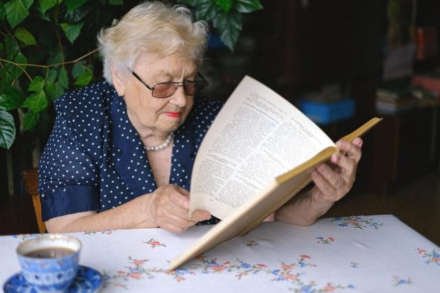 An older adult woman sits at a table with a cup of tea and reads a book.