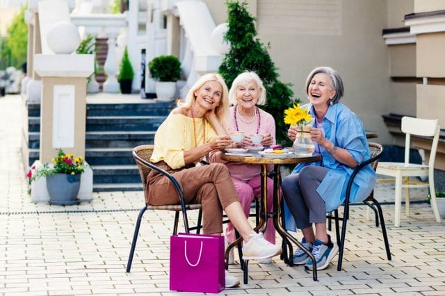 Three older adult women sit at an outdoor cafe table smiling and drinking tea. There is a shopping bag on the ground.