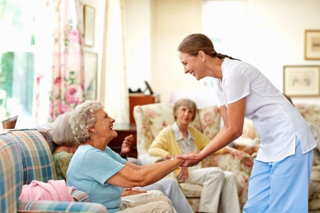 A young woman wearing scrubs stands and holds both hands of an older adult woman who is sitting on a sofa. They both smile. Another older adult woman sitting in an armchair looks at them.