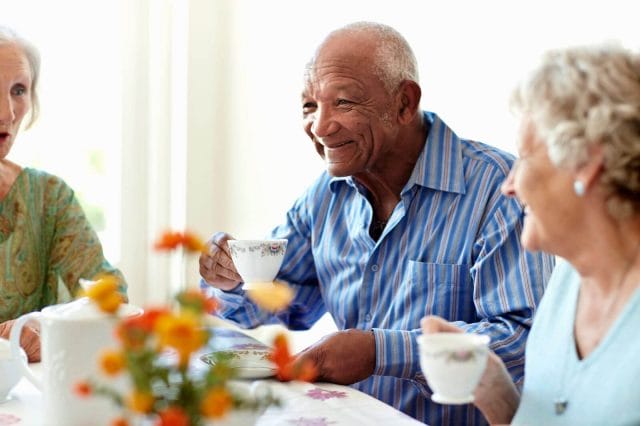 An older adult man sits at a table drinking tea with his two older adult women friends.