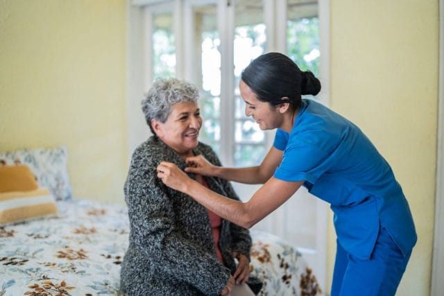 A young woman wearing scrubs helps an older adult woman, who is sitting on a bed, put on her sweater.