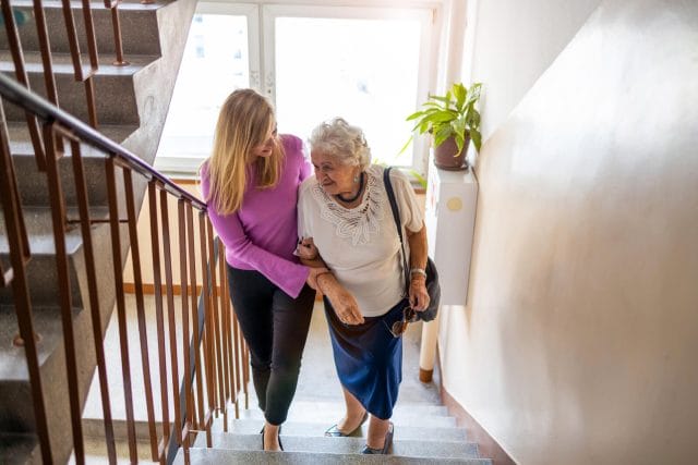 A youthful woman helps an older adult woman walk up a flight of stairs.
