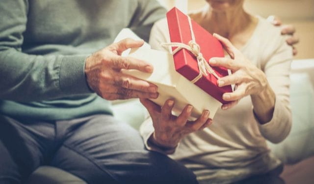 An older adult man holds a gift for an older adult woman as she opens it.