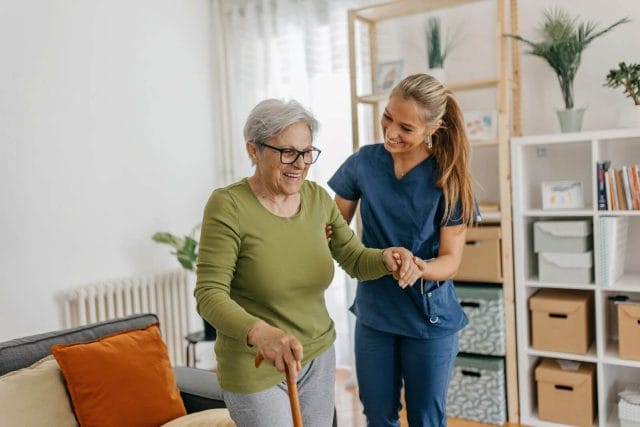An older adult woman with a cane stands with the assistance of a younger adult woman wearing scrubs.