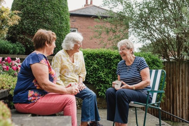 Three older adult women sit outside smiling and drinking coffee.
