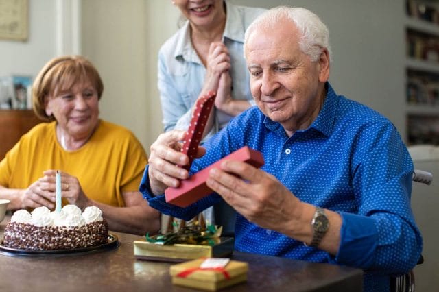 An older adult man opens a gift as two women watch, smiling.