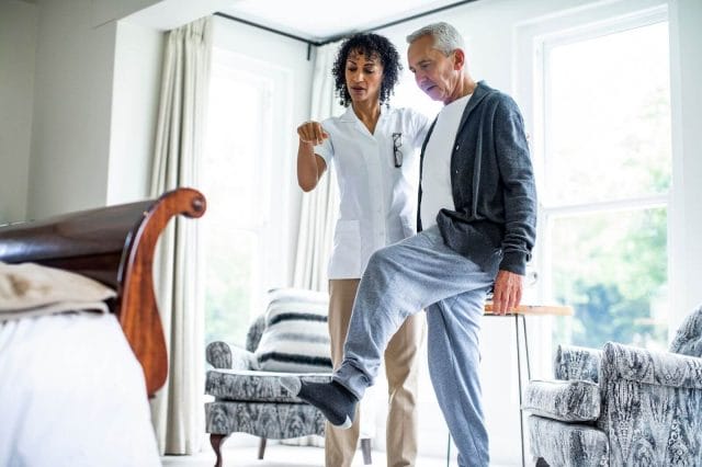 An older adult man stands on one leg while a younger woman doctor assists him.